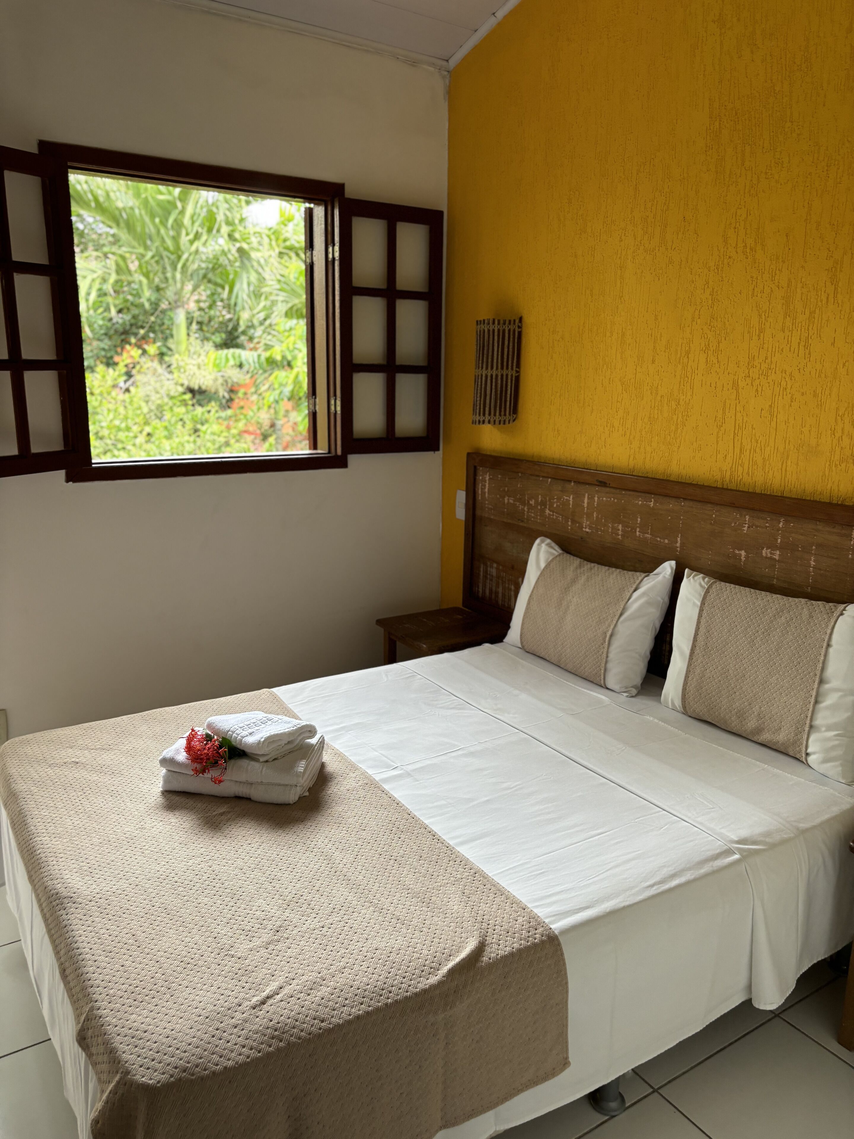 Una cama bien tendida con una colcha color beige y almohadas, una mesita de noche con una lámpara, y una ventana con vista a la vegetación.