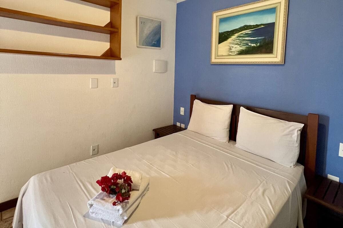 Un lit bien fait, avec du linge de lit blanc et un bouquet de fleurs rouges, un tableau représentant un paysage côtier et un plafond à rayures bleues et blanches.