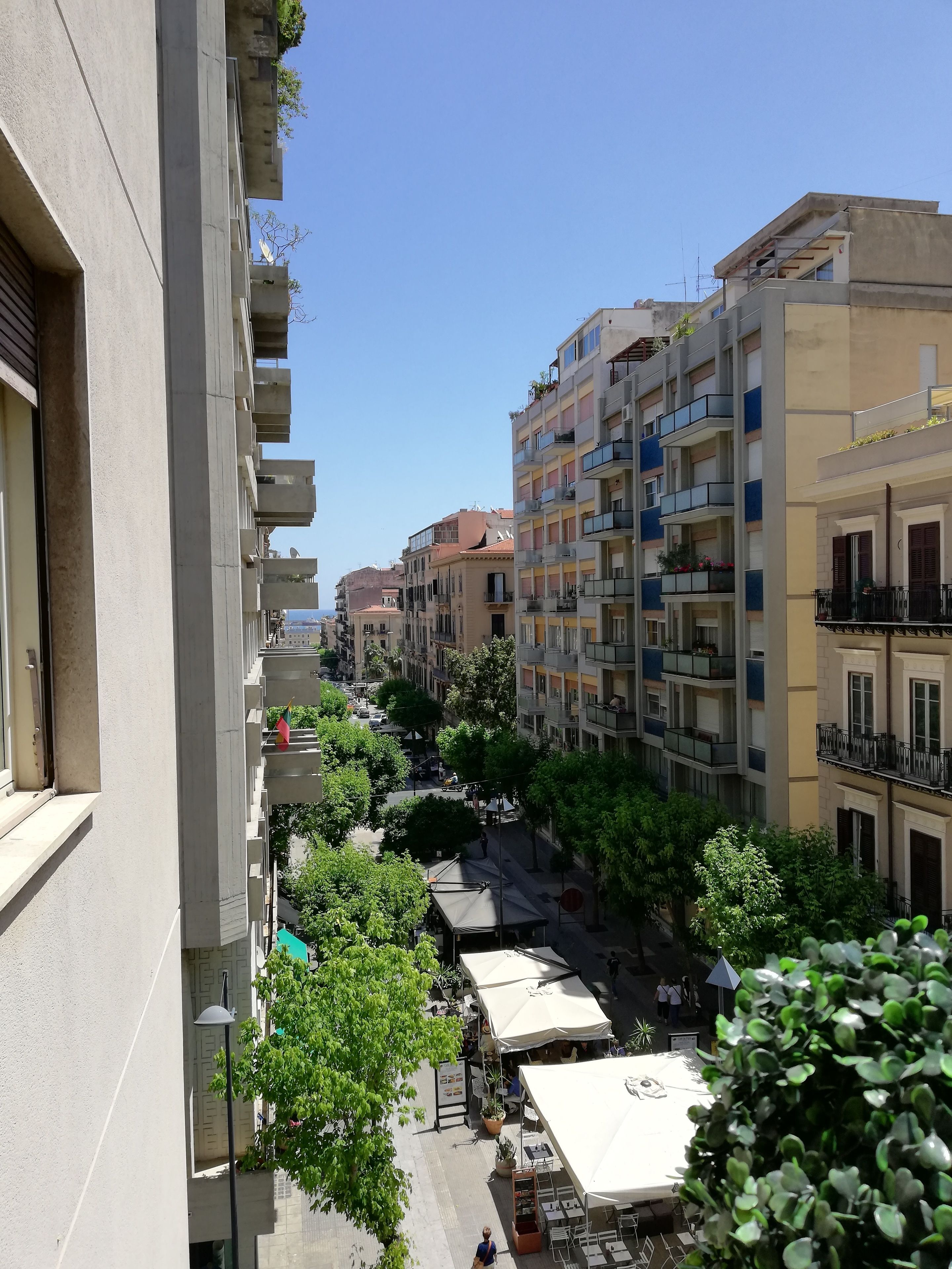 A multi-story residential building with balconies, a street with shops, and trees.