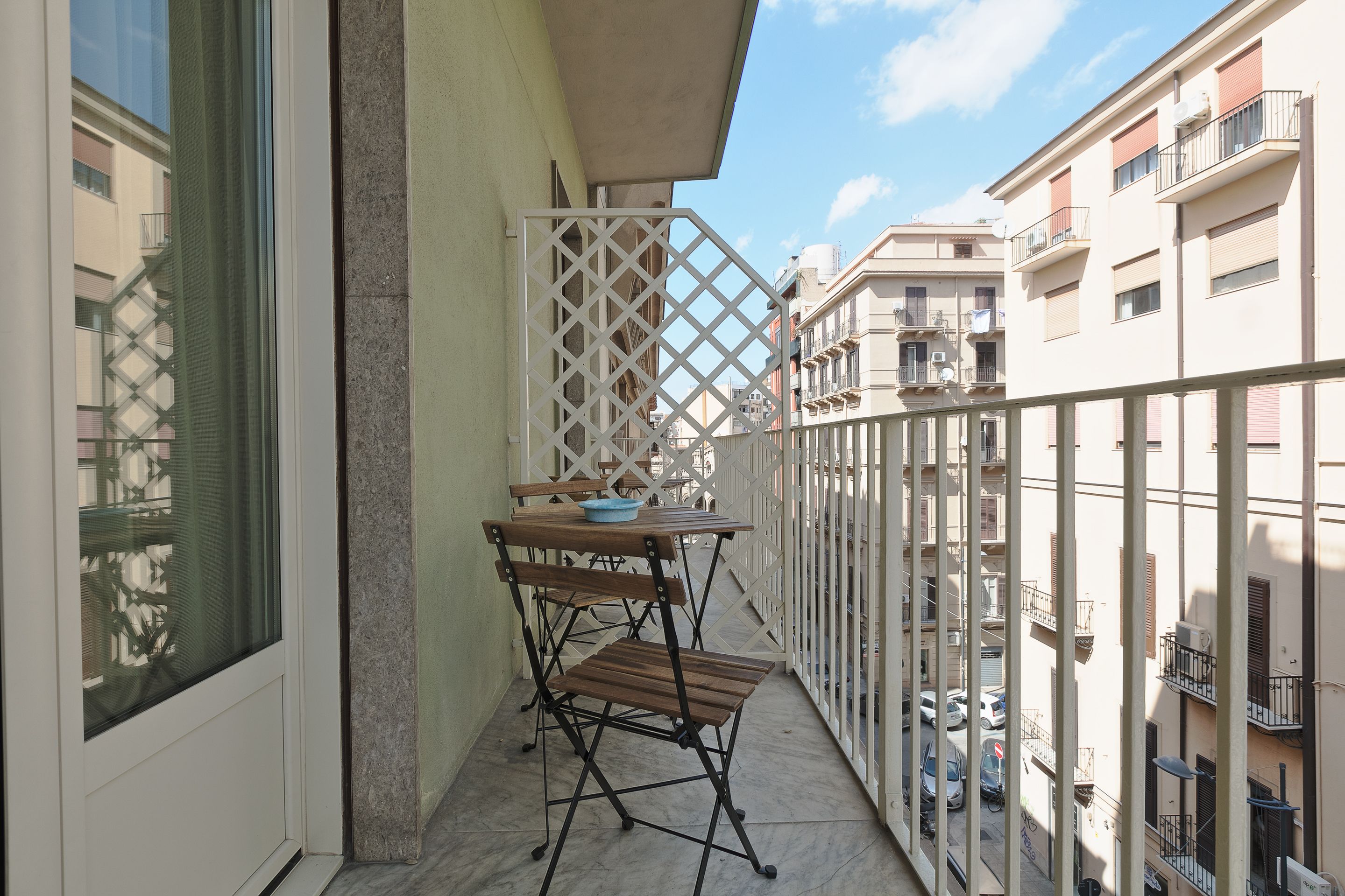 Un balcón con una mesa y sillas, un panel de celosía blanca y vistas a edificios cercanos.
