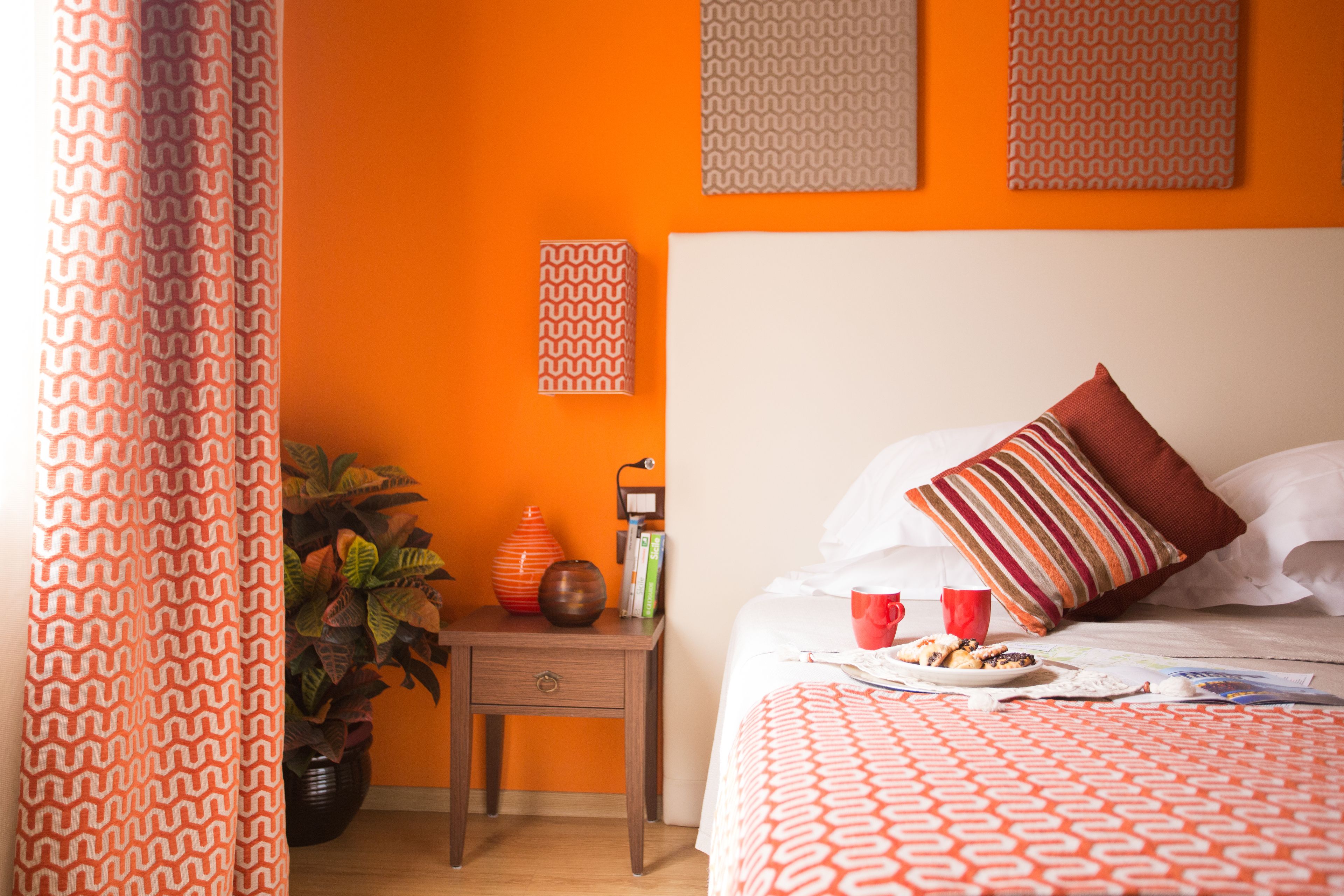 Une chambre avec un mur orange, une table de chevet en bois ornée d’un vase et de livres, un lit recouvert d’une couvre-lit à motifs et un oreiller à rayures.