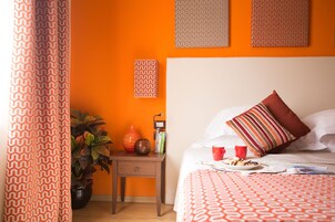 A bedroom with an orange wall, a wooden nightstand with a vase and books, a bed with a patterned bedspread, and a striped pillow.