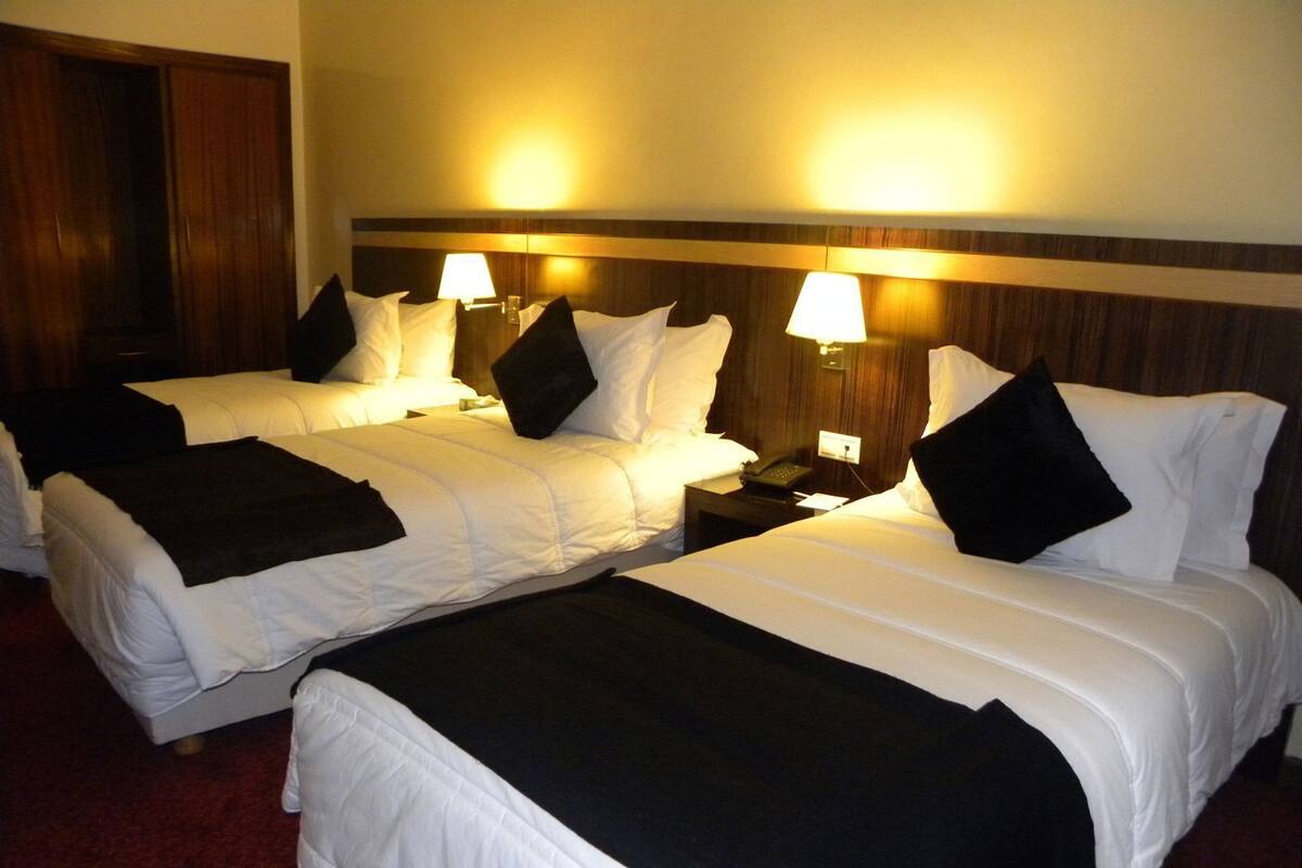 A hotel room with two beds, each with white bedding and black throw pillows, a wooden headboard, bedside lamps, and a red carpet.