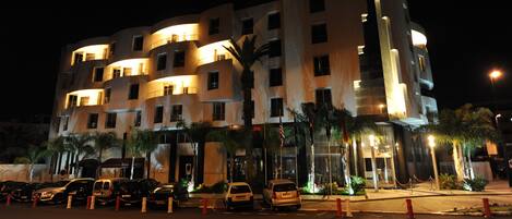 A multi-story hotel with a prominent "RIVERE HOTEL" sign, a parking lot with several cars, and a well-lit entrance.