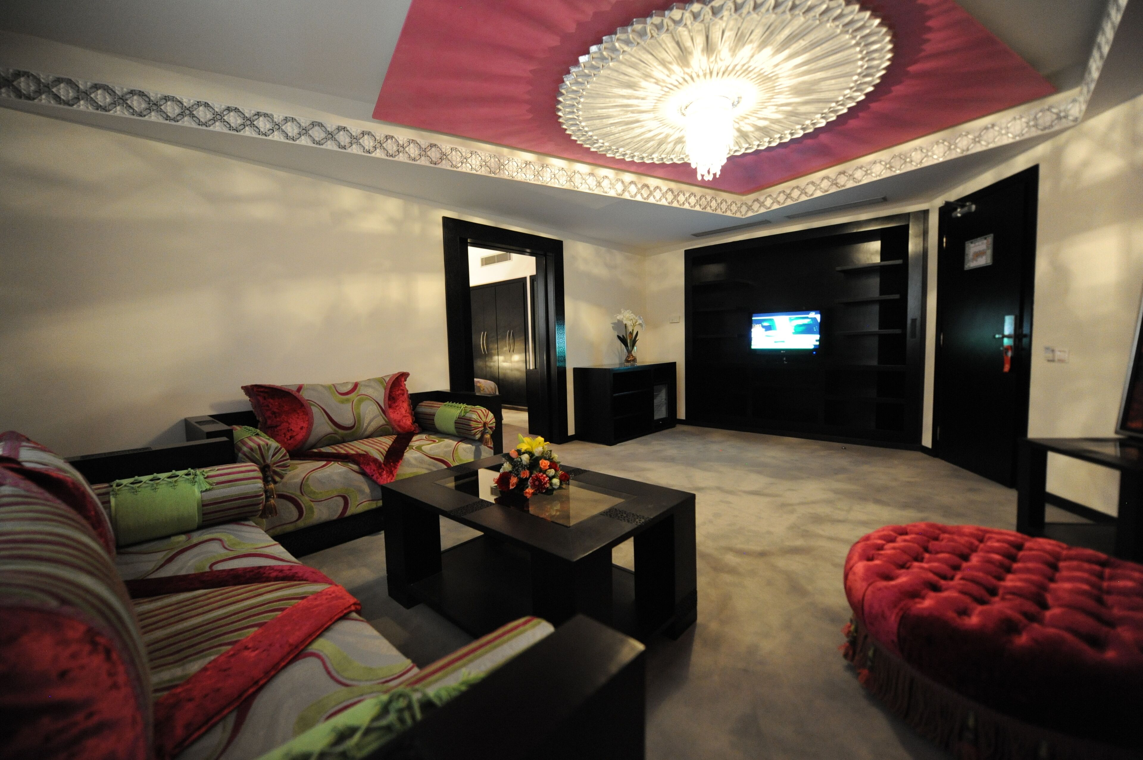 A living room with a sofa, a coffee table with flowers, a red ottoman, and a TV in the background.