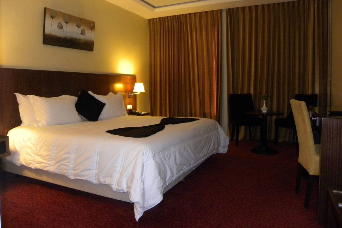 A hotel room with a large bed, a nightstand with a lamp, a chair, a table, and a painting on the wall.