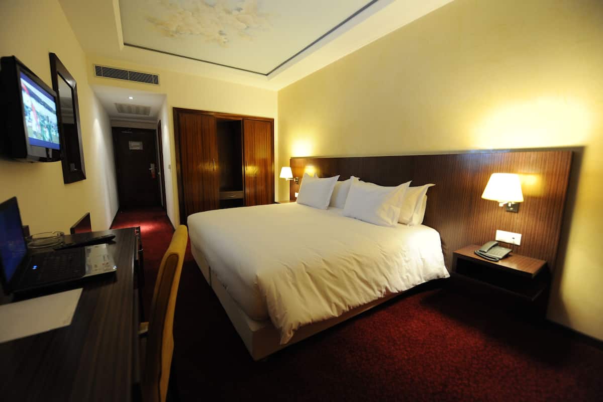 A hotel room with a large bed, a desk with a laptop, a TV, and a nightstand with a lamp.
