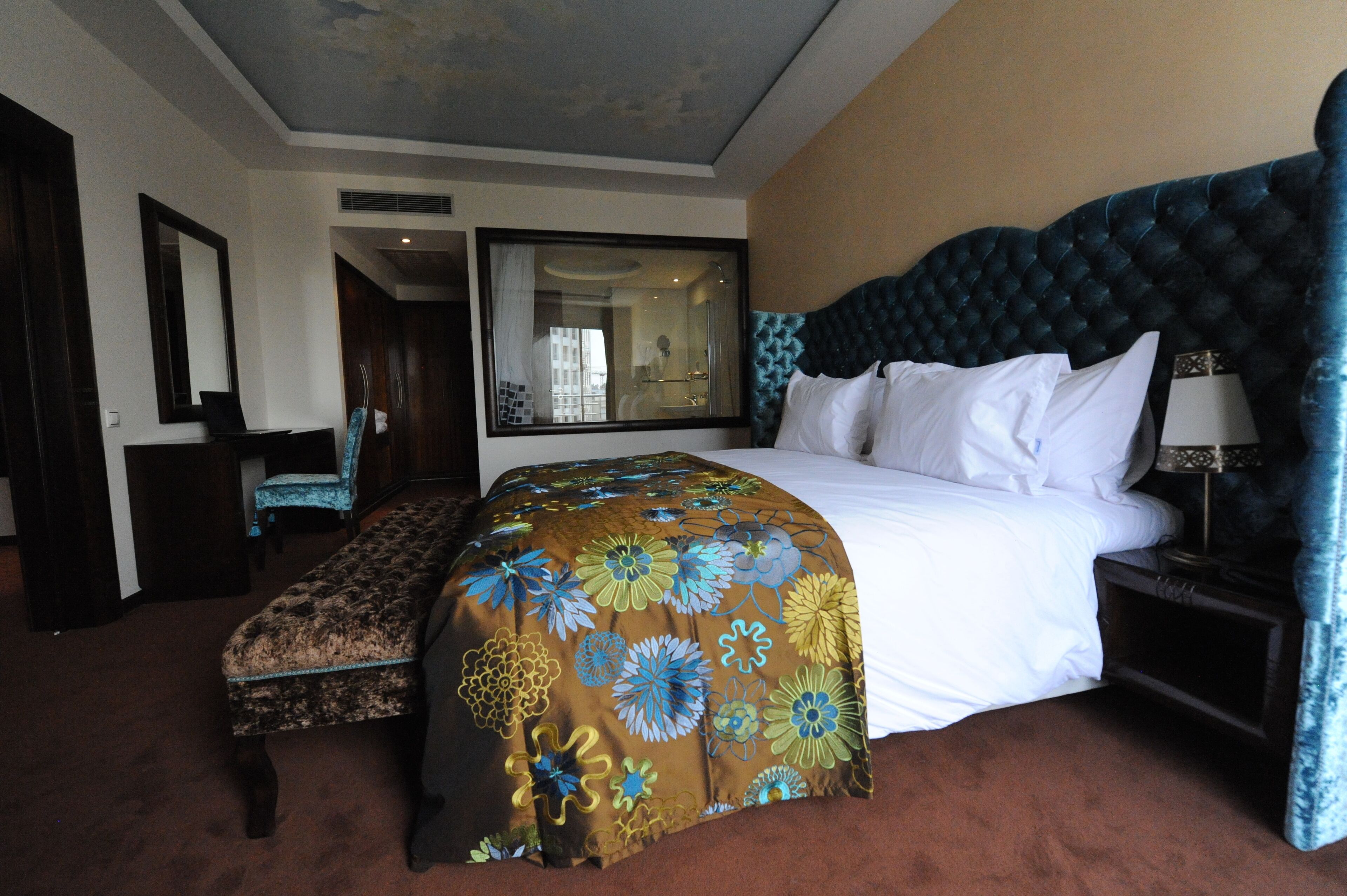 A hotel room with a large bed, a bench, a chair, a mirror, and a nightstand with a lamp.