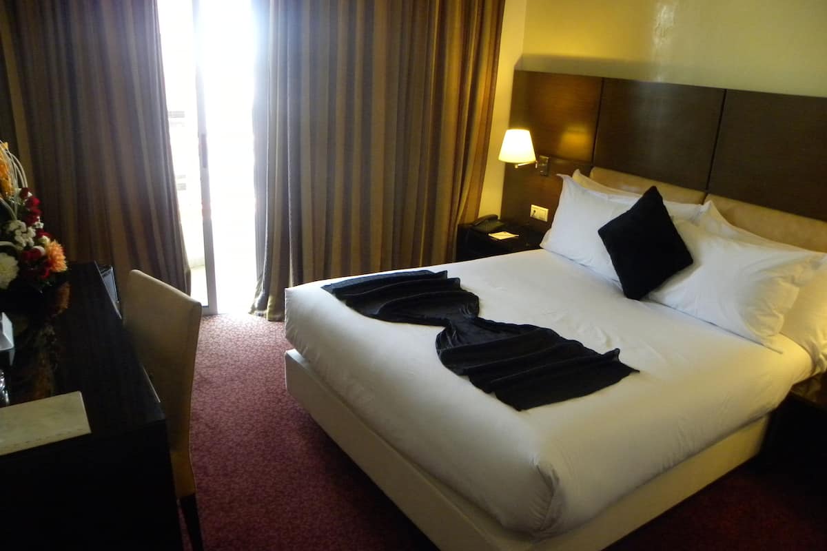 A hotel room with a large bed, a bedside table with a lamp, a chair, and a desk with a vase of flowers.