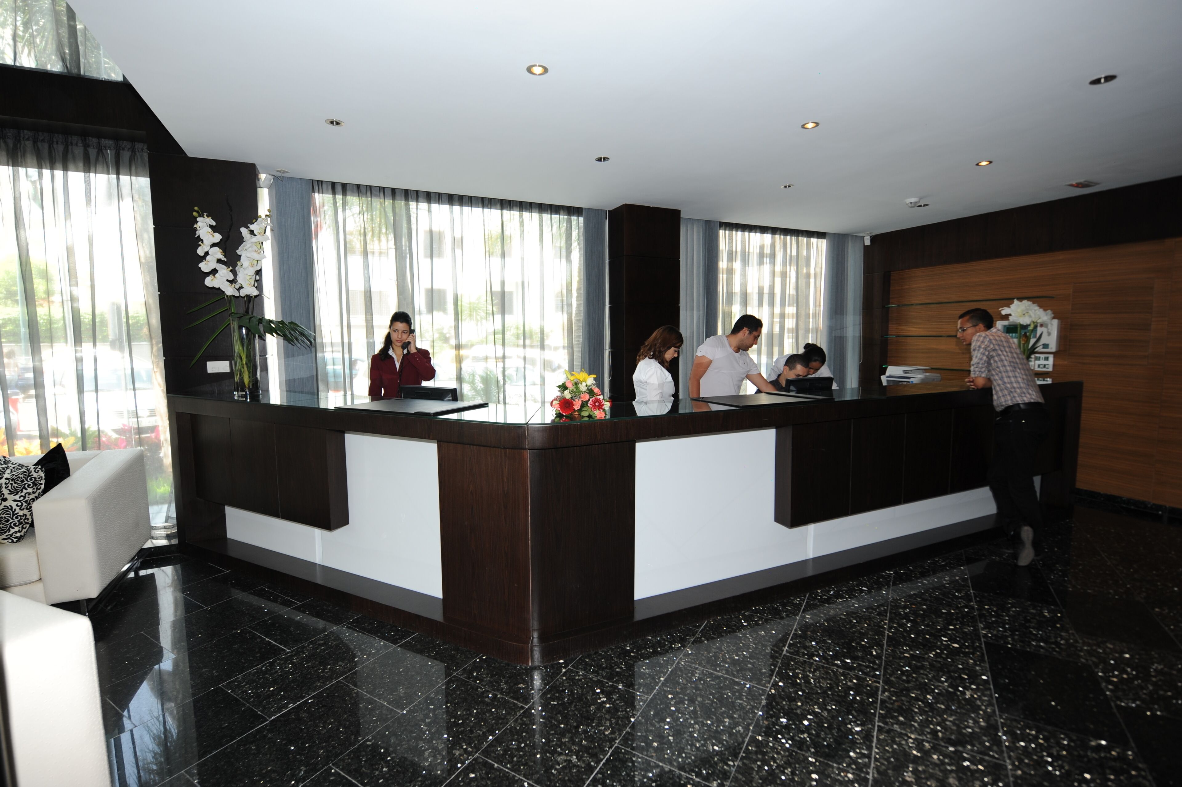 A modern hotel reception area with a dark wood reception desk, white walls, and a large window offering a view of the outdoors.