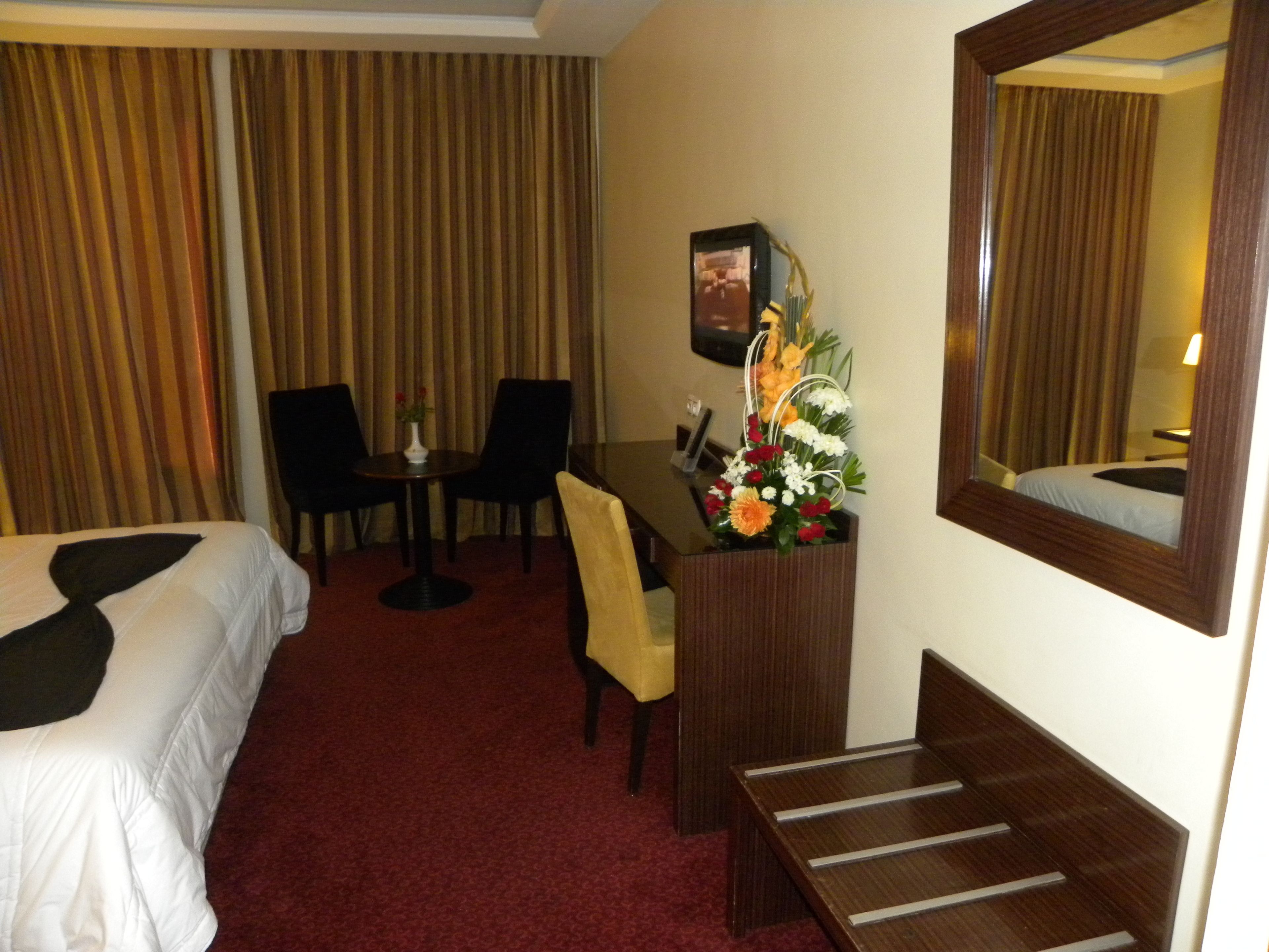 A hotel room with a bed, a desk with a chair, a small table with a vase of flowers, and a mirror on the wall.