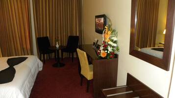 A hotel room with a bed, a desk with a chair, a small table with a vase of flowers, and a mirror on the wall.