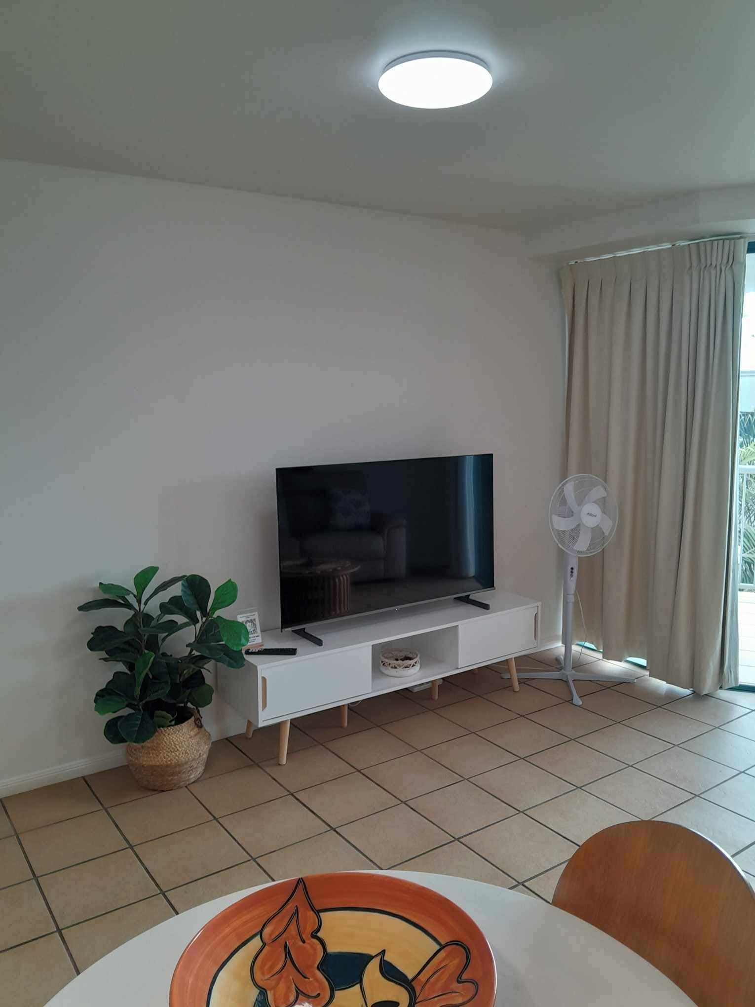 A living room with a flat-screen TV on a white TV stand, a potted plant, a standing fan, and a dining area with a table and chairs.