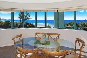 A round glass table with wooden chairs in a room with large windows overlooking a beach.