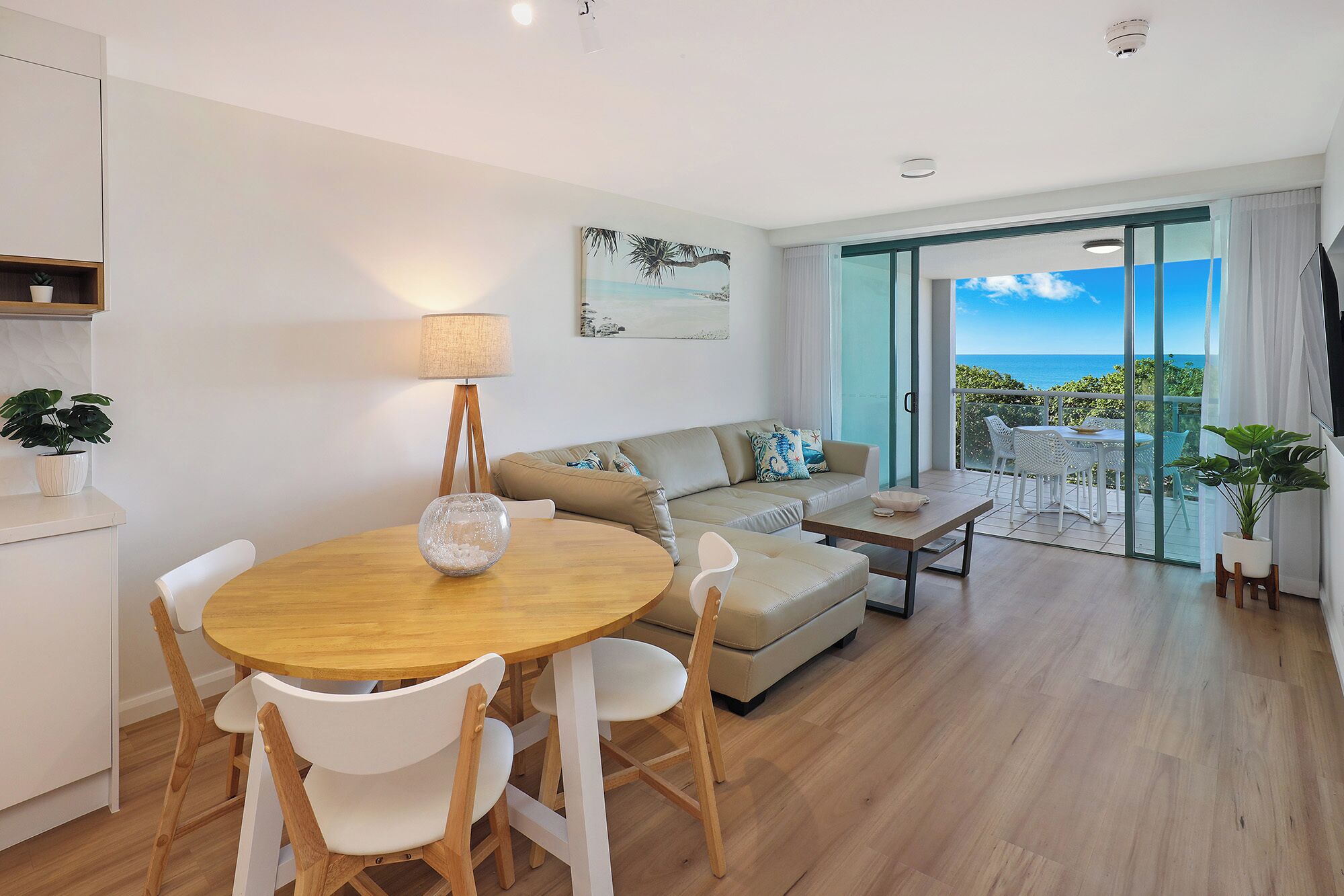 A modern living room with a round wooden table, white chairs, a beige sofa, and a balcony with a view of the sea.