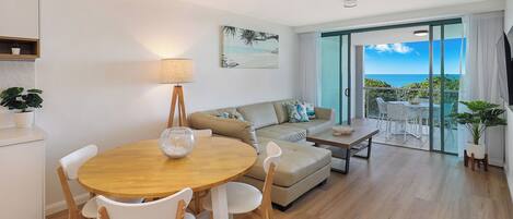 A modern living room with a round wooden table, white chairs, a beige sofa, and a balcony with a view of the sea.