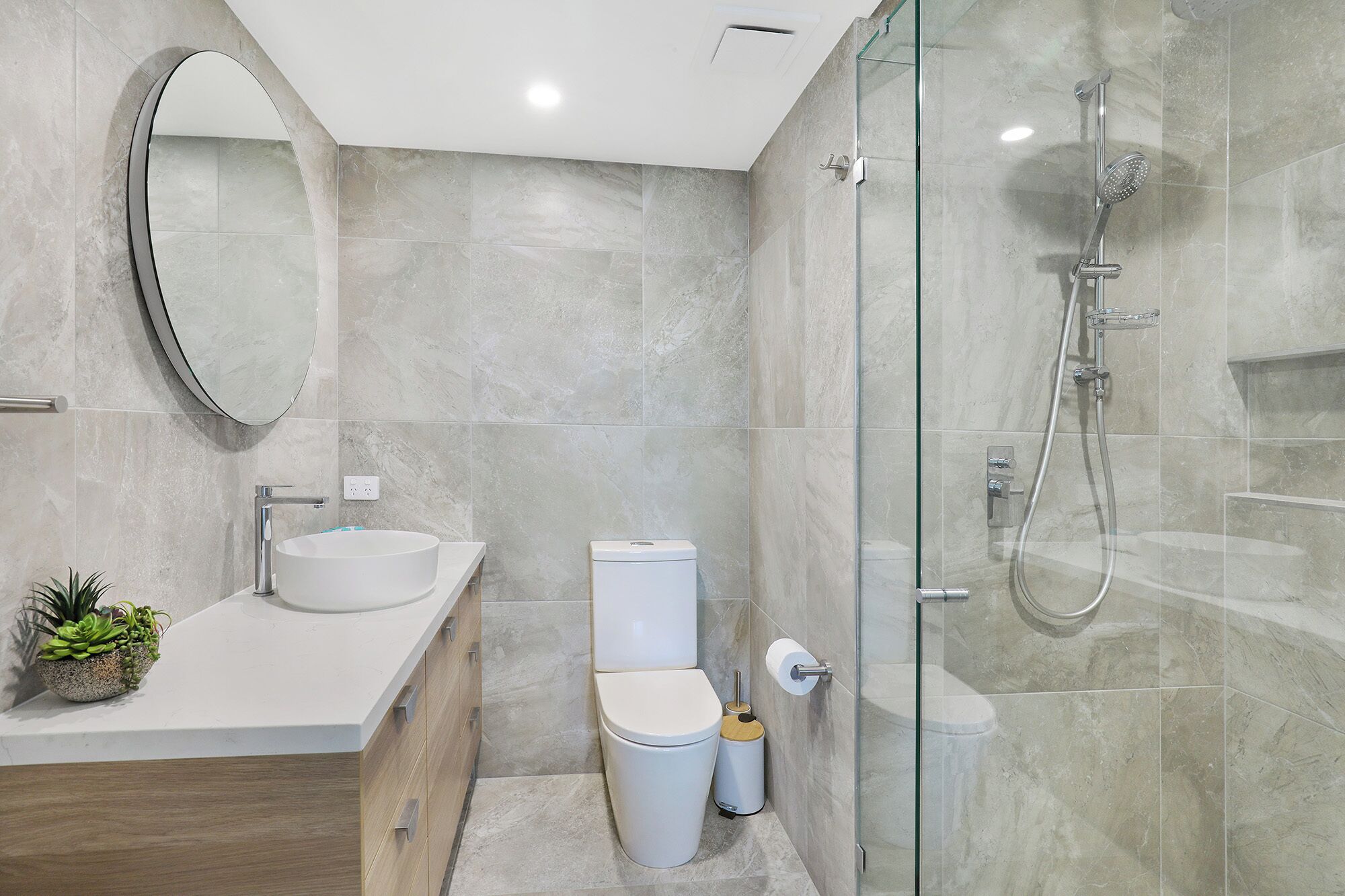 Un baño moderno con cabina de ducha de vidrio, un espejo redondo, un lavamanos y un inodoro.