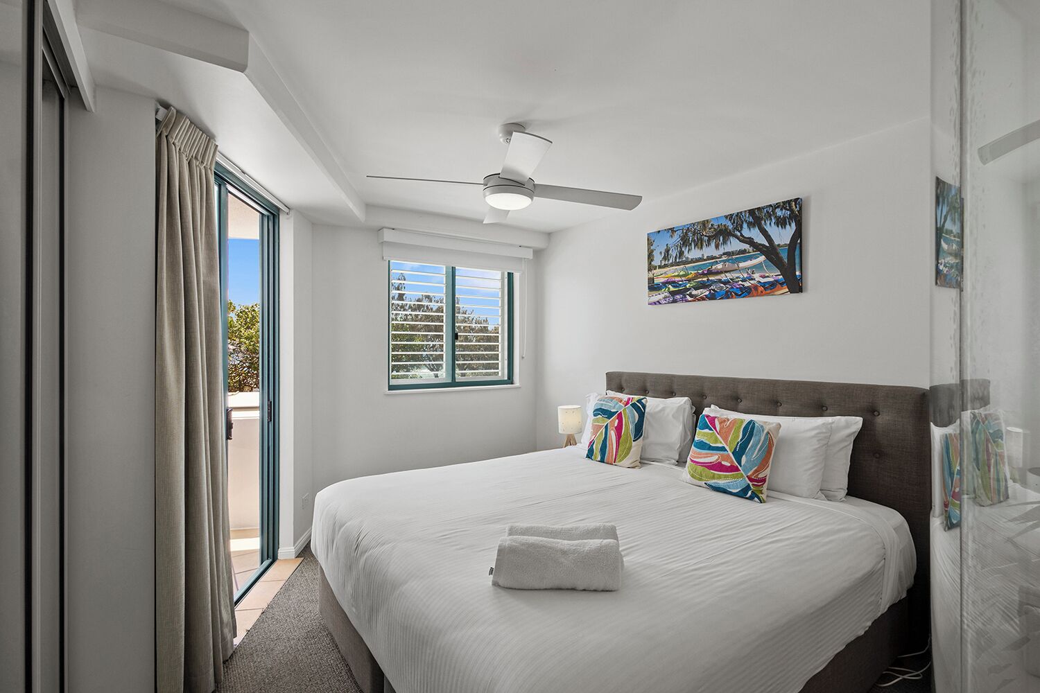 A bedroom with a large bed, a ceiling fan, a window with blinds, and a painting on the wall.