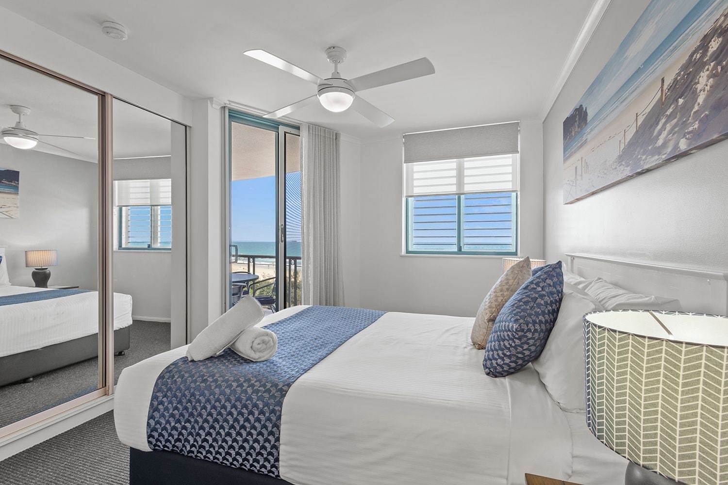 A hotel room with a bed, a ceiling fan, a window with blinds, and a view of the beach.