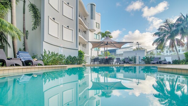 Zona de piscina con sillas de descanso y sombrillas, rodeada de palmeras y un edificio con balcones.