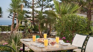 Mesa al aire libre con alimentos para el desayuno y bebidas, rodeada de plantas tropicales y vista al mar.