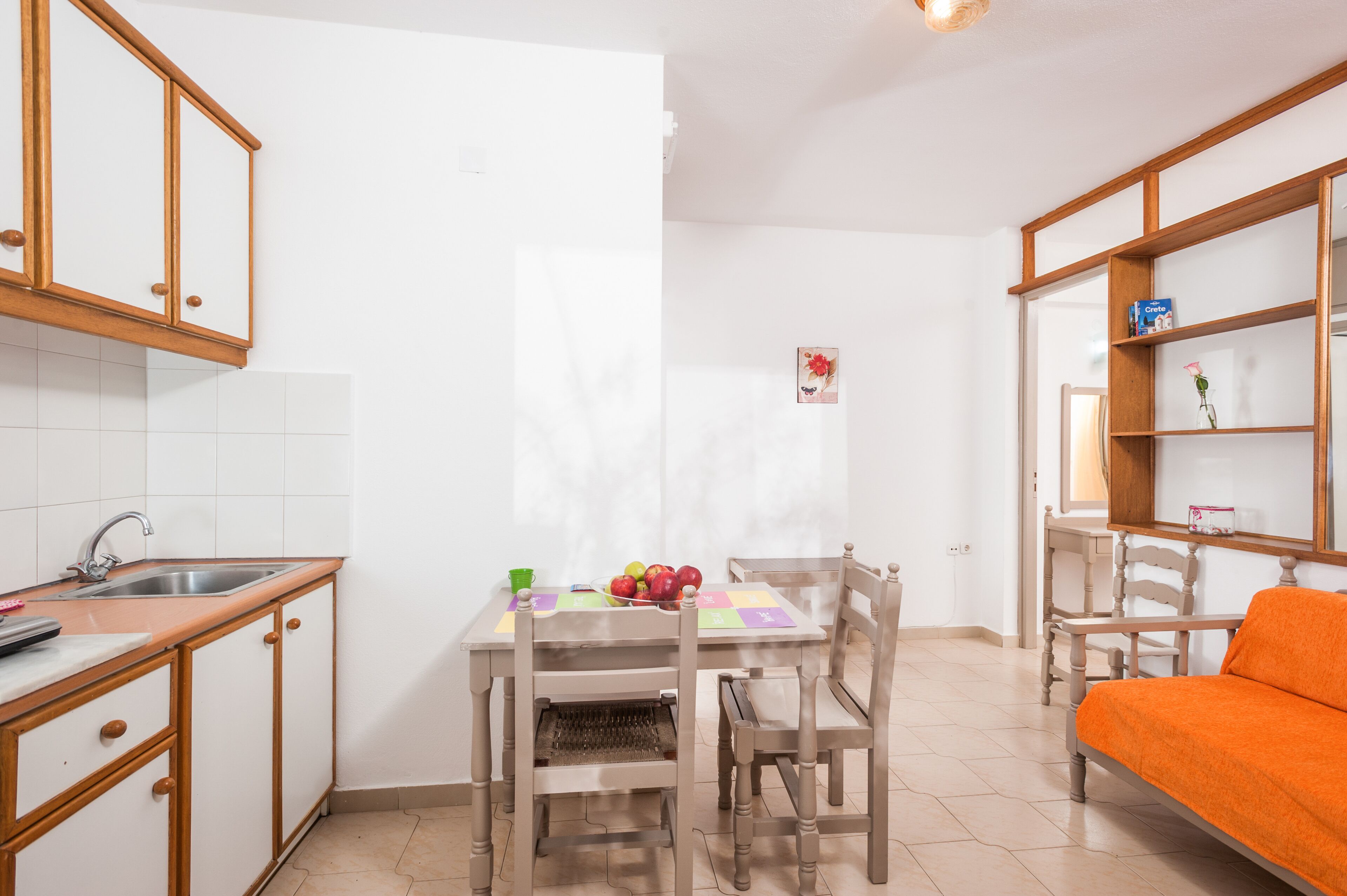 Una piccola cucina con lavello, pensili in legno, un tavolo da pranzo con sedie e un letto con coperta arancione.