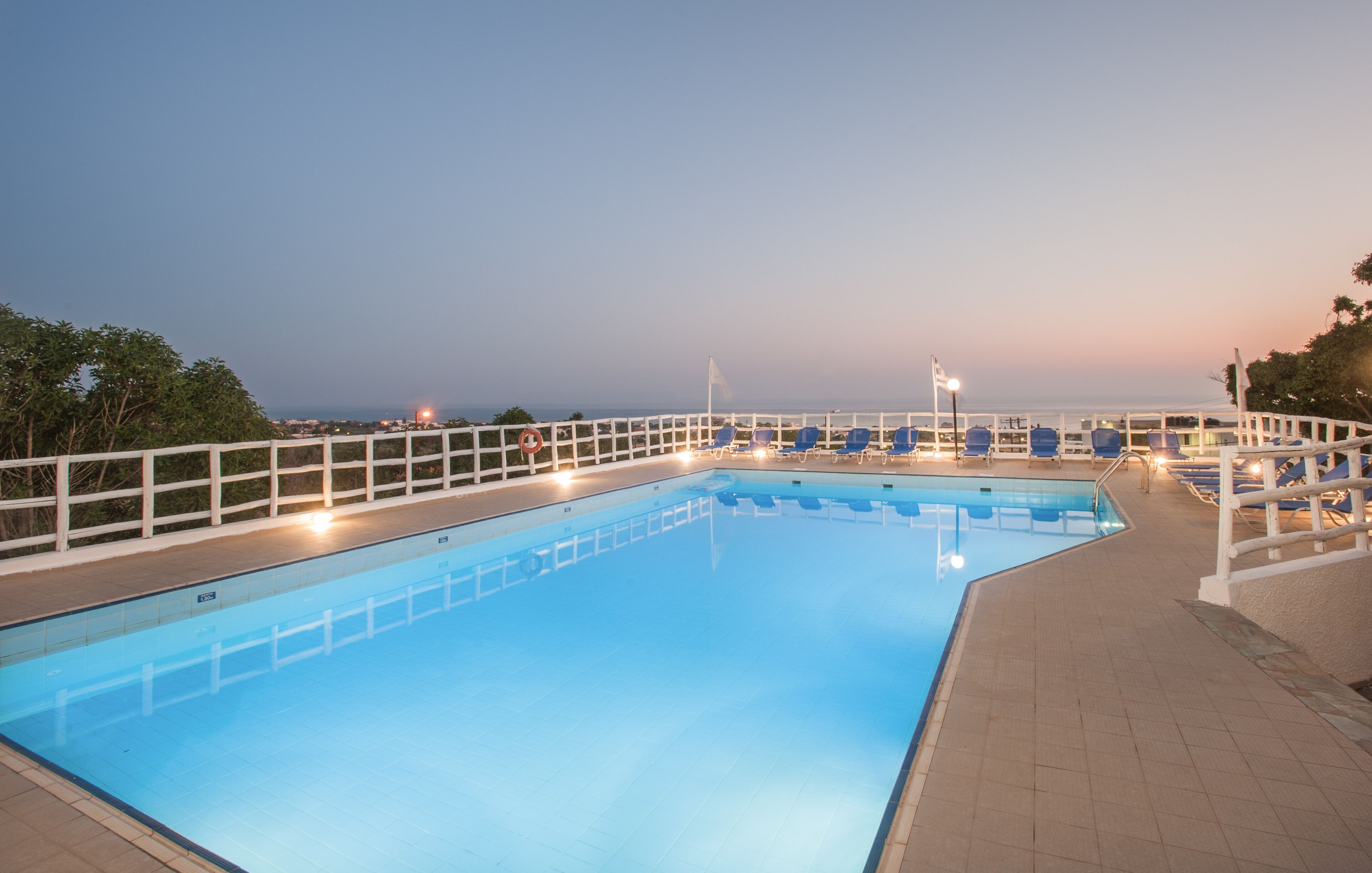 A swimming pool with a white fence, blue lounge chairs, and a view of the sea at dusk.