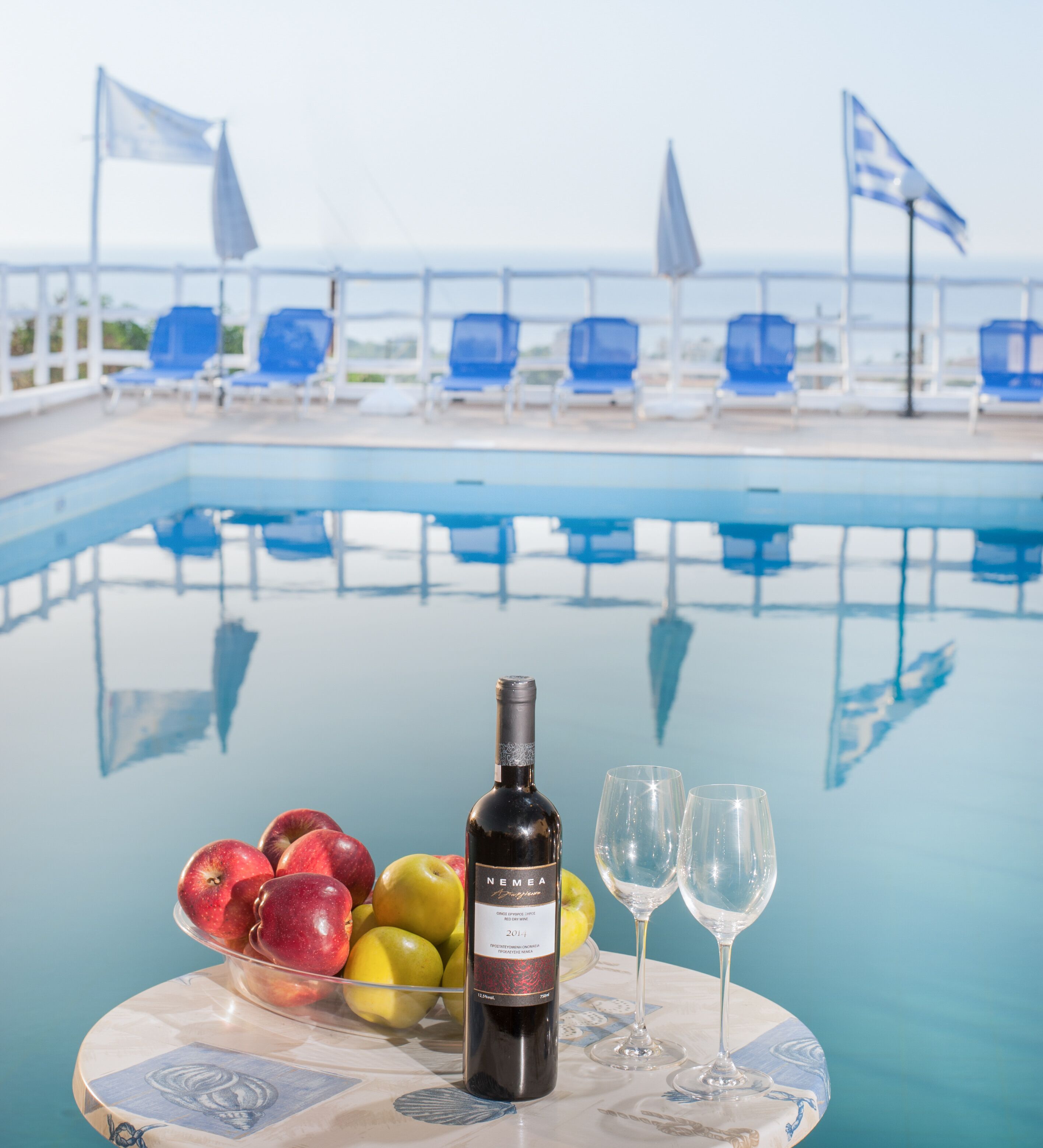 Mesa junto a la piscina con una botella de vino, dos copas y un bol de manzanas.
