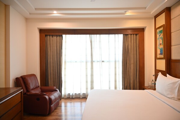 A hotel room with a bed, a leather chair, a wooden dresser, and a large window with curtains.