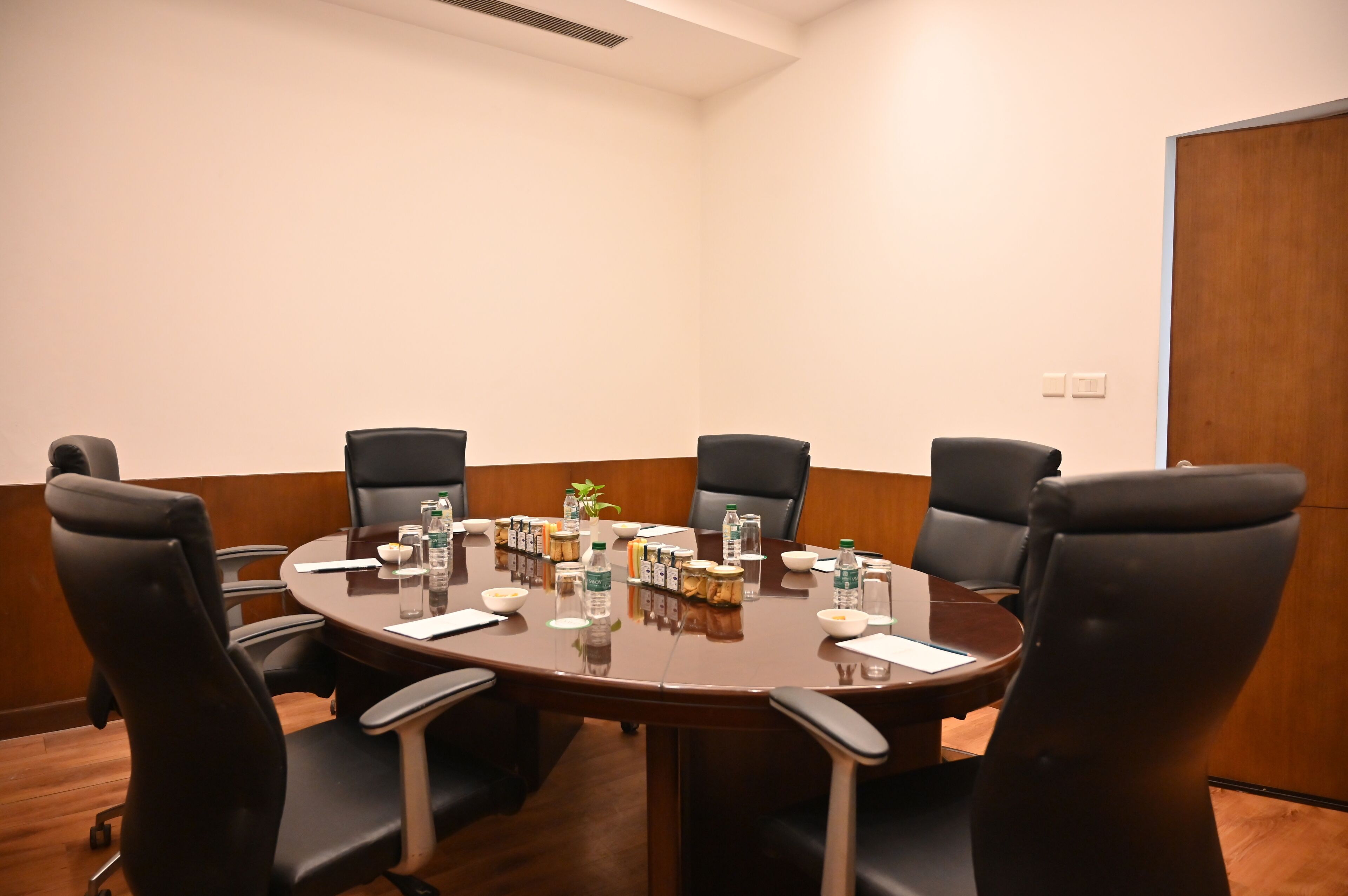 Una sala de conferencias con una mesa redonda, seis sillas negras y varios objetos como botellas de agua, cuencos y frascos sobre la mesa.