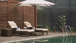 A poolside area with lounge chairs and an umbrella, surrounded by potted plants and a glass wall. - Savoy Suites Greater Noida (Greater Noida)