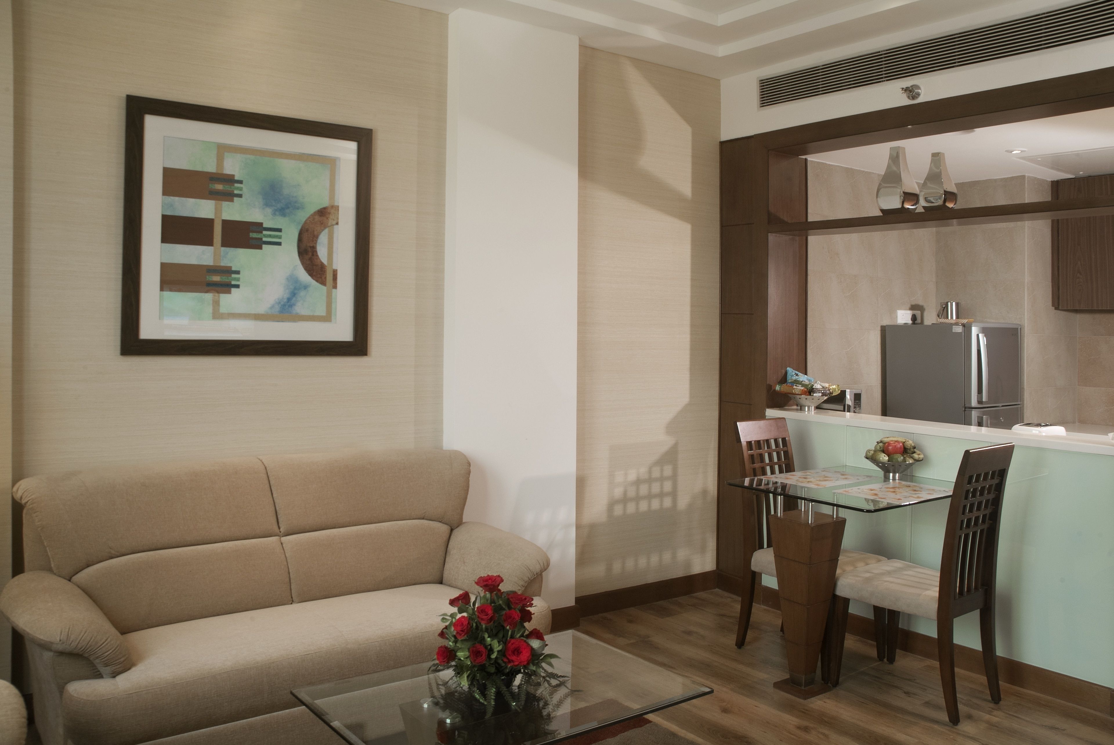 A living room with a sofa, a glass coffee table with a vase of flowers, a dining area with a table and chairs, and a kitchen area with a refrigerator and cabinets.