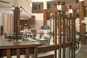 A spacious hotel lobby with multiple dining areas, large columns, and decorative lighting fixtures.