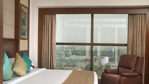 A hotel room with a large bed, a brown leather chair, and a view of a cityscape through large windows. - Savoy Suites Greater Noida (Greater Noida)