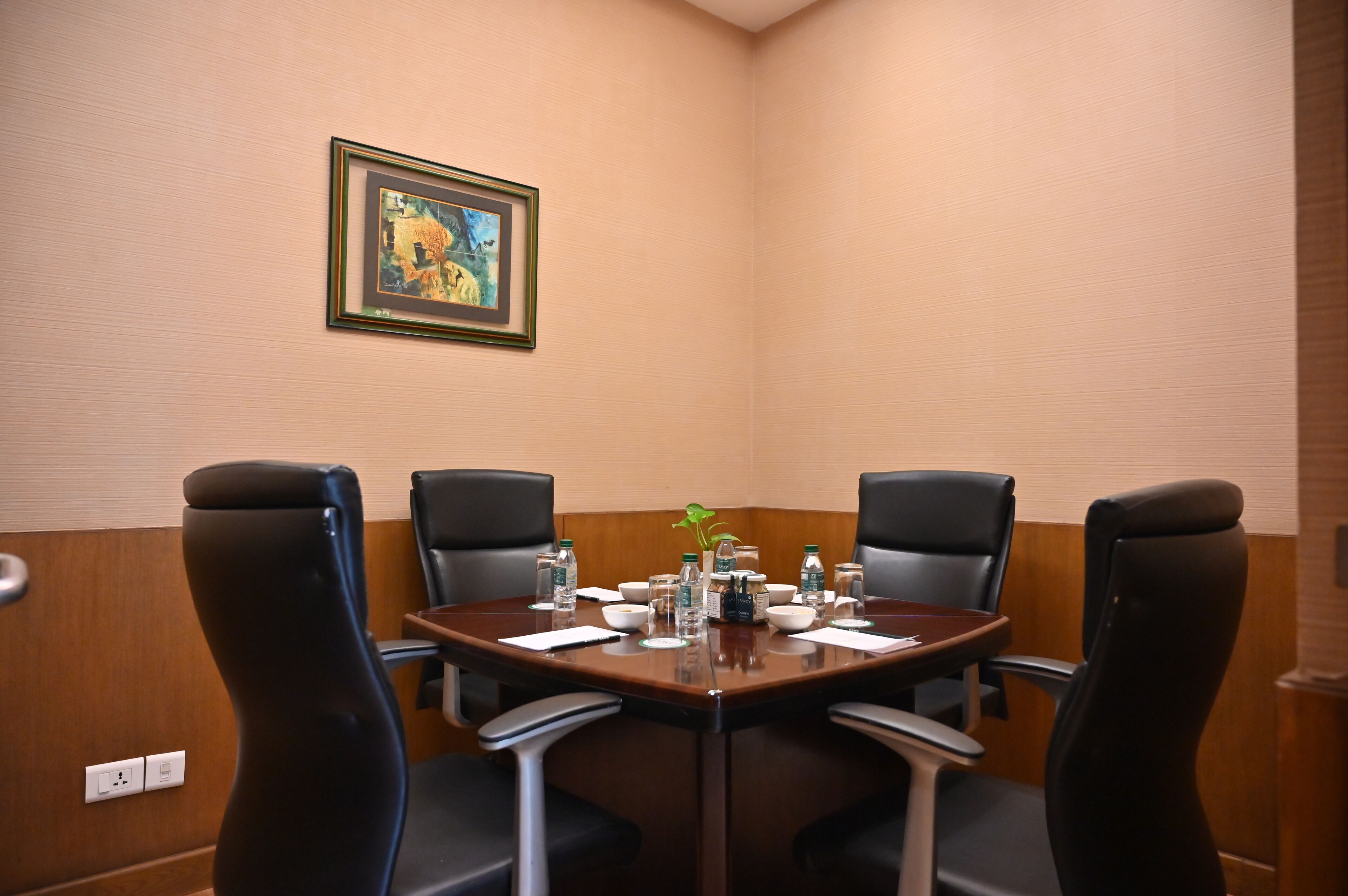A conference room with a round table, four chairs, a painting on the wall, and various items on the table including water bottles and a plant.