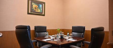 A conference room with a round table, four chairs, a painting on the wall, and various items on the table including water bottles and a plant.