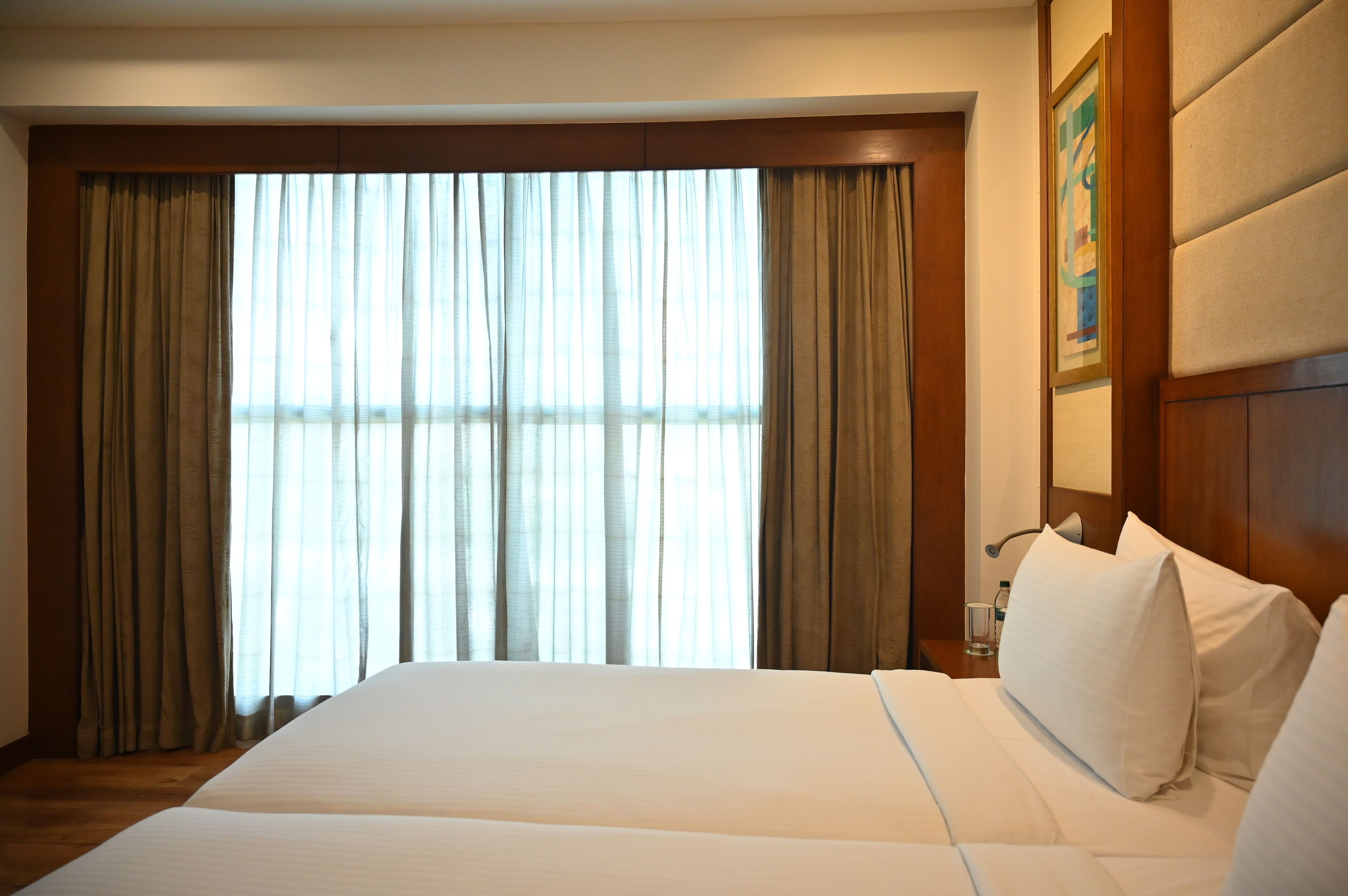 A hotel room with a large bed, a wooden headboard, a nightstand with a glass and a bottle, and a window with curtains.