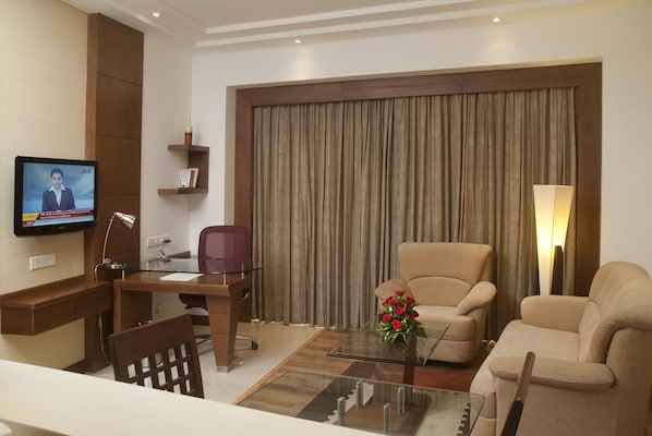 Una habitación de hotel moderna con escritorio, dos sillas, sofá, televisor y un ventanal con cortinas.