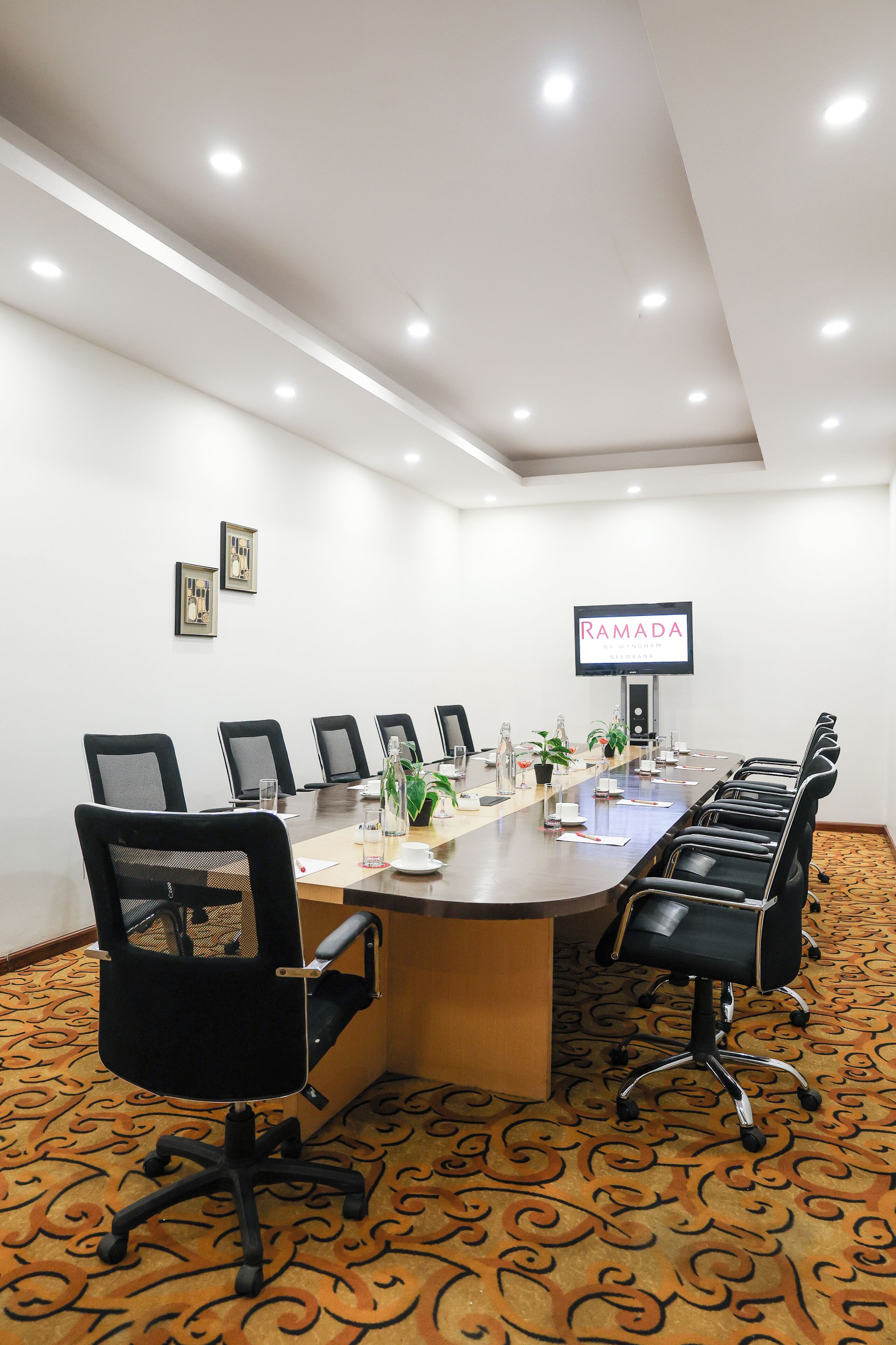 Uma sala de conferências com uma mesa comprida, cadeiras, uma tela de TV e piso coberto de carpete.
