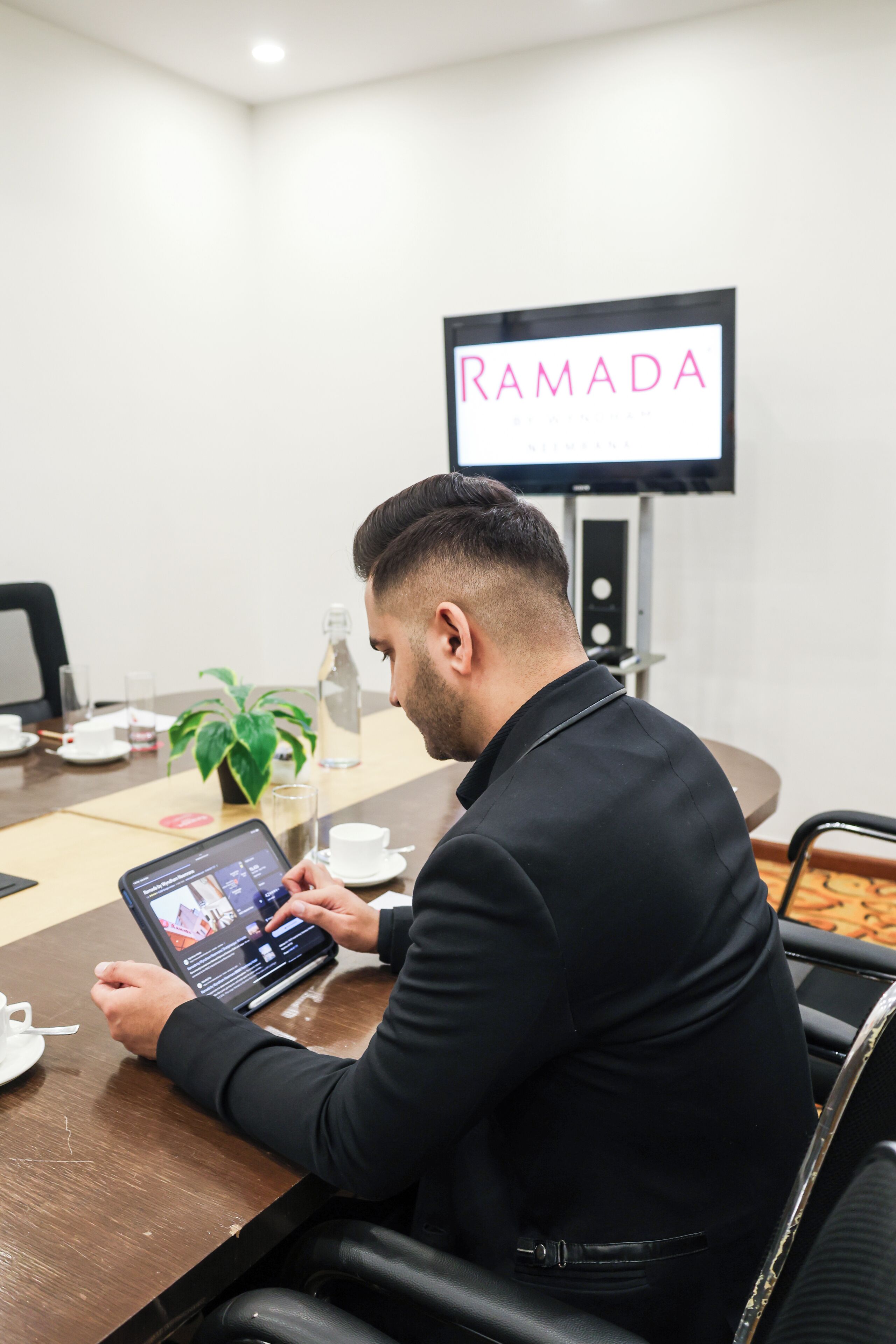 Um homem sentado em uma mesa de conferência com um laptop, uma planta e uma garrafa de água.