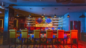 A bar area with a colorful tiled wall, multiple hanging pendant lights, and a bar stocked with bottles. There are several bar stools with cushioned seats. - Ramada by Wyndham Neemrana (Neemrana)