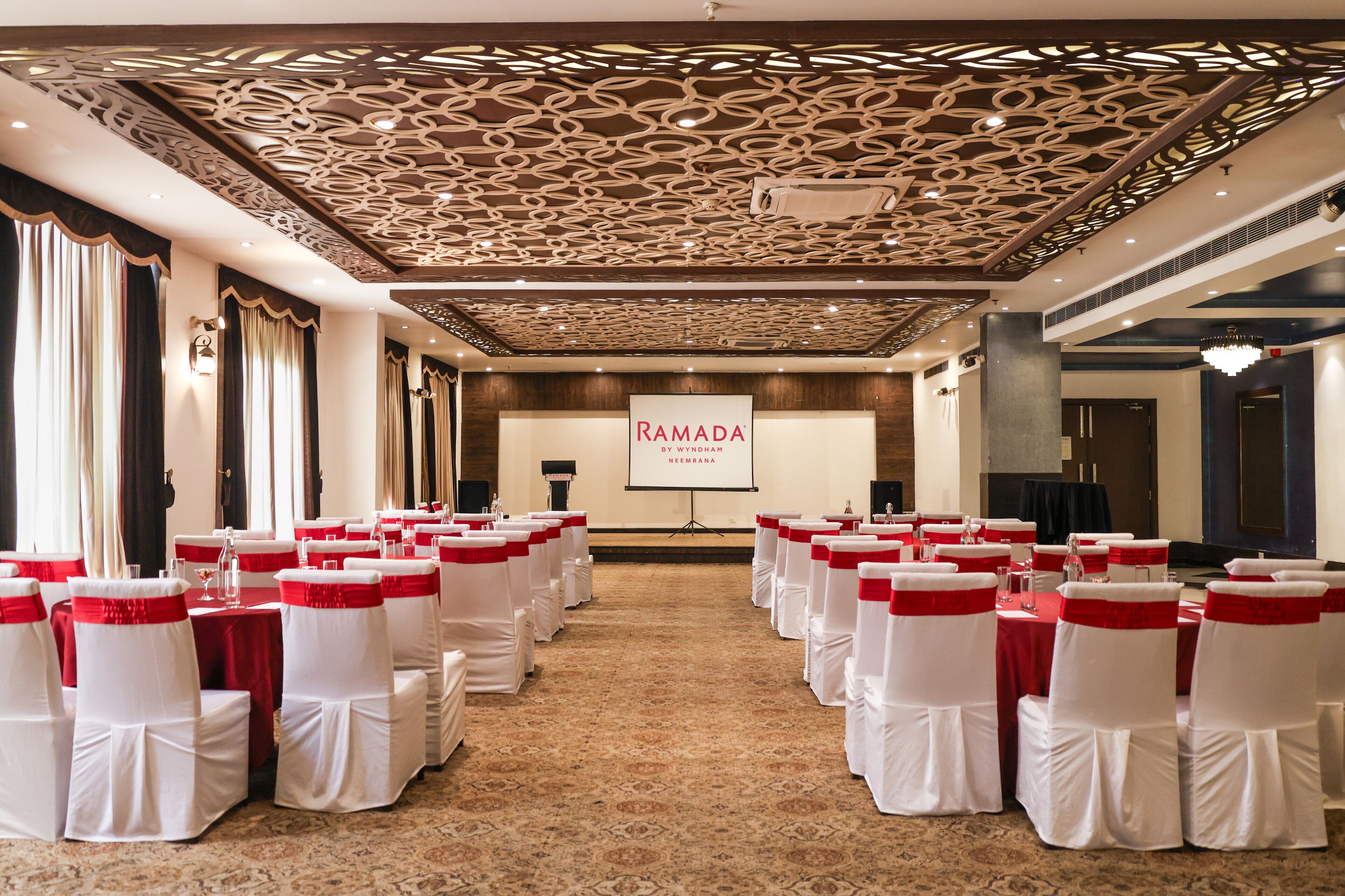 Une salle de conférence avec des rangées de chaises blanches ornées de rubans rouges, un écran de projection et une enseigne « RAMADA ».