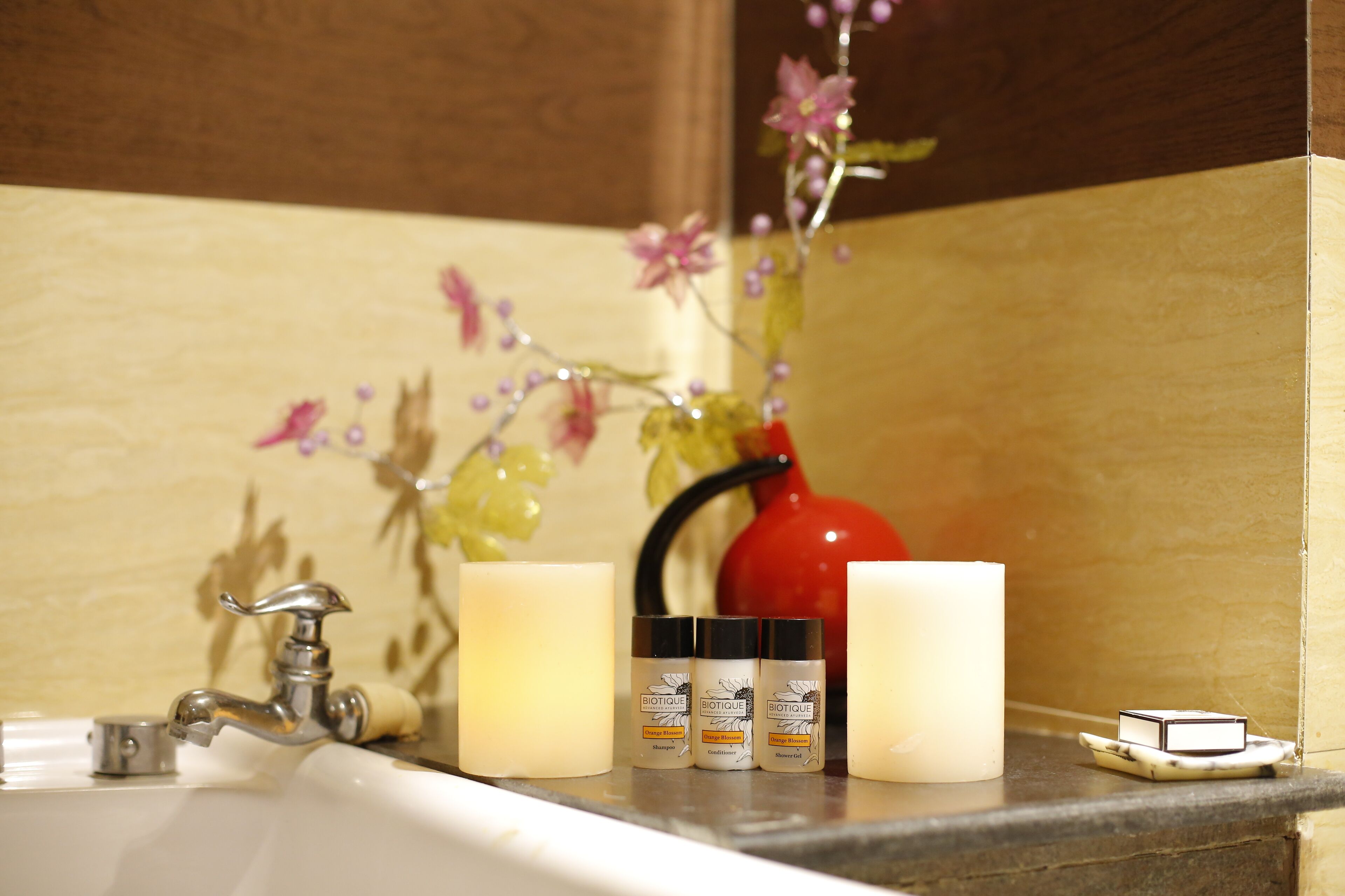 A bathroom sink with a faucet, two candles, a red vase with flowers, and three Biotique skincare bottles.