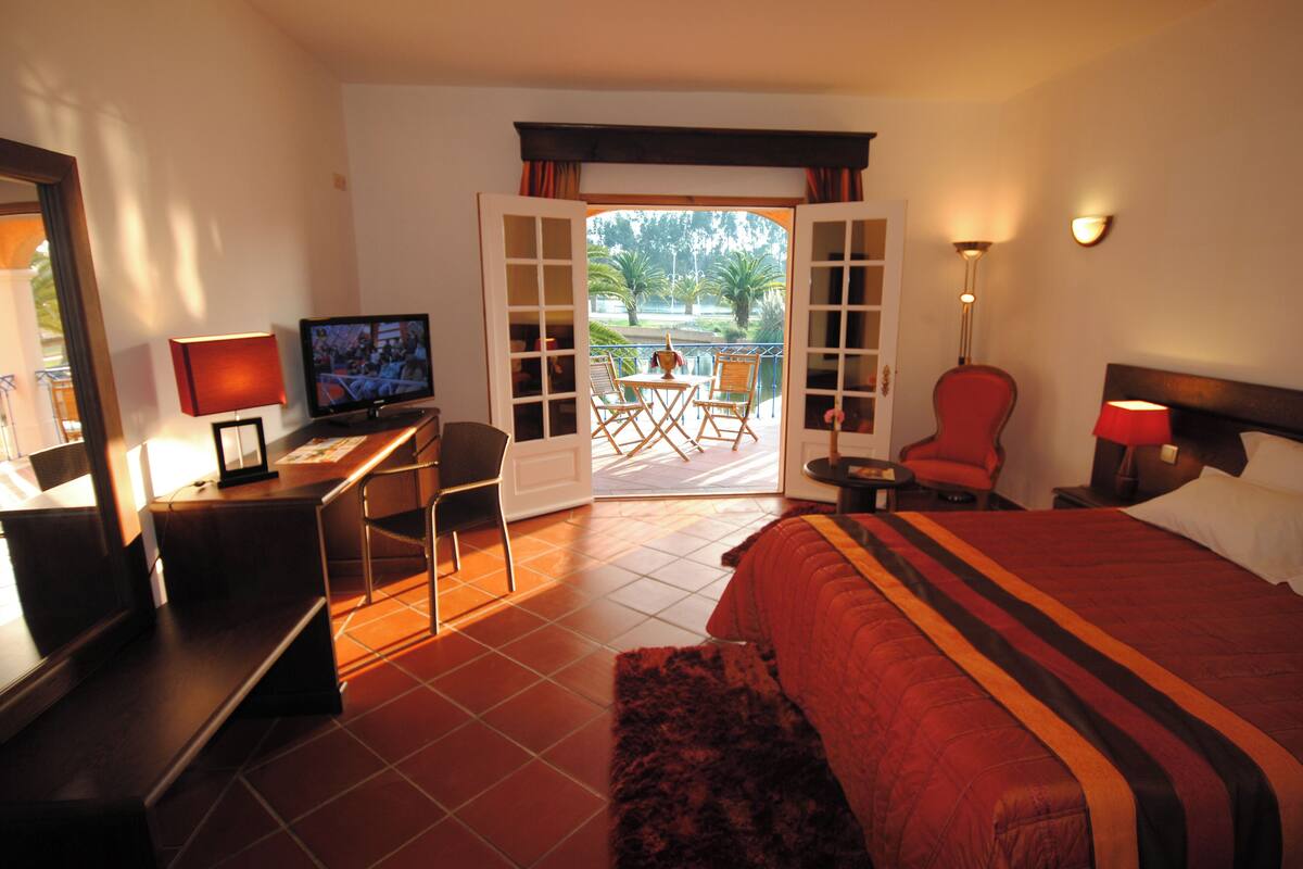A hotel room with a bed, a desk with a TV, a chair, and a view of a balcony with outdoor furniture.