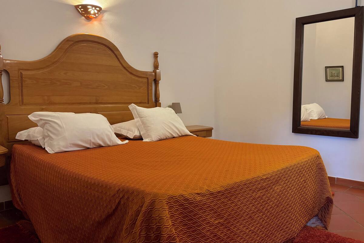A neatly made bed with a wooden headboard, white pillows, and an orange bedspread. There is a nightstand with a lamp and a wall-mounted mirror reflecting part of the room.