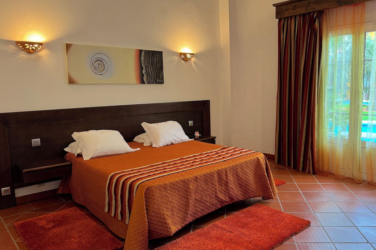 A hotel room with a large bed, two bedside tables, a nightstand, a wall-mounted artwork, and a view of a swimming pool through curtains.
