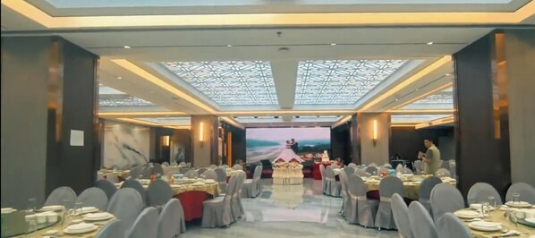 Un salón de banquetes con mesas redondas, sillas y un escenario con fondo decorativo.