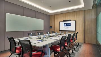 Una sala de conferencias con una mesa larga, sillas, una pizarra, una pantalla mostrando el logotipo de Holiday Inn Express y un dispensador de agua.