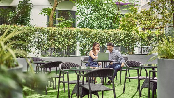 Un espacio al aire libre con sillas y mesas de mimbre, rodeado de follaje y un edificio al fondo.