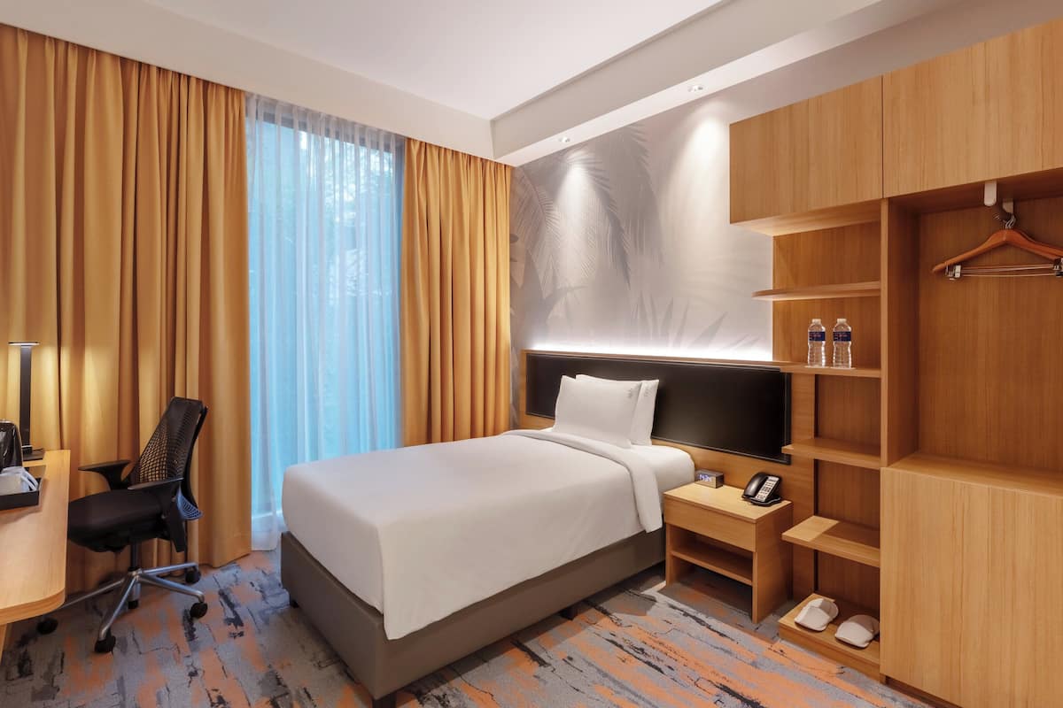 A hotel room with a bed, desk, chair, TV, bedside table with phone and lamp, wardrobe, and a window with curtains.